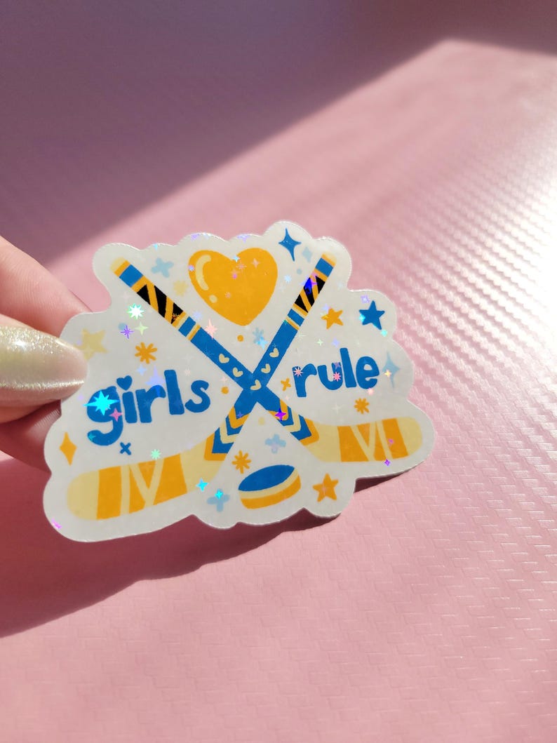 May include: A holographic sticker with the words "Girls rule" and hockey-themed graphics. The design features crossed hockey sticks, a puck, a heart, and stars in blue, yellow, and white. The sticker has a glittery, iridescent finish.