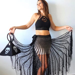 May include: A black crochet halter top paired with a matching black crochet skirt featuring an open-weave fringe design. A black pyramid-shaped crochet handbag with white skull motifs and a circular handle is held to the side of the outfit.