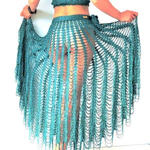 May include: A dark teal crochet skirt featuring an open-work mesh pattern with long, dangling fringe strands that create a layered, flowing effect. The skirt has a fitted waistband and is crafted from a shimmering, metallic-toned yarn material.