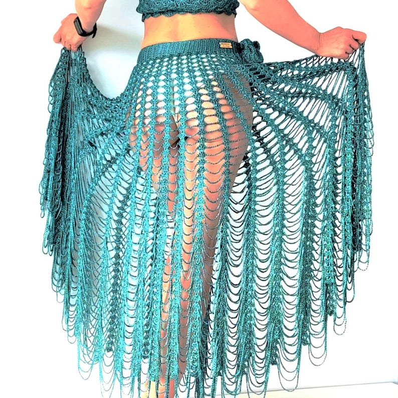 May include: A dark teal crochet skirt featuring an open-work mesh pattern with long, dangling fringe strands that create a layered, flowing effect. The skirt has a fitted waistband and is crafted from a shimmering, metallic-toned yarn material.