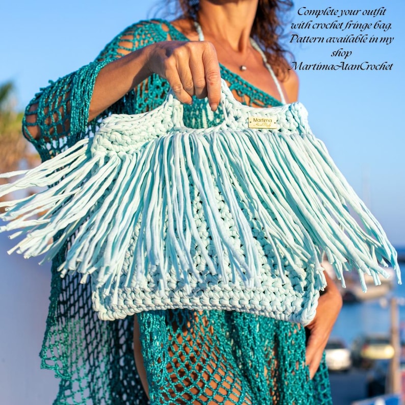 May include: A light blue crochet handbag featuring long fringe detailing and a small rectangular tag with the text MartimaAtanCrochet. The bag is constructed with a thick yarn texture and includes two integrated handles at the top of the structure.