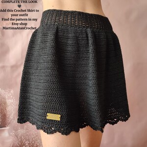May include: A black crochet skirt featuring a wide, open-weave waistband and a solid, textured body with a scalloped hem. A small rectangular gold-colored tag with the text Martima is attached to the lower front of the skirt.
