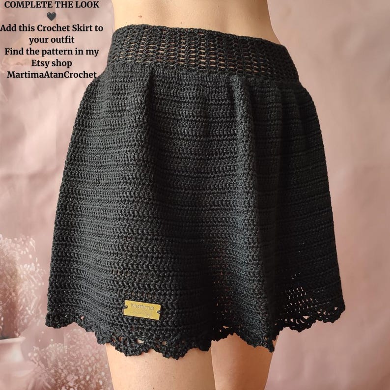May include: A black crochet skirt featuring a wide, open-weave waistband and a solid, textured body with a scalloped hem. A small rectangular gold-colored tag with the text Martima is attached to the lower front of the skirt.