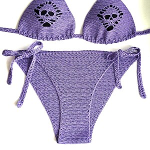 K&ouml;nnte beinhalten: Ein lavendelfarbenes H&auml;kel-Bikini-Set bestehend aus einem Triangel-Oberteil mit Totenkopf-Ausschnitt und einer passenden Bikinihose zum Binden an den Seiten. Der Stoff ist in einem strukturierten Maschenmuster gefertigt.