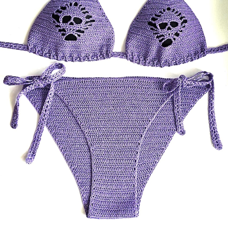K&ouml;nnte beinhalten: Ein lavendelfarbenes H&auml;kel-Bikini-Set bestehend aus einem Triangel-Oberteil mit Totenkopf-Ausschnitt und einer passenden Bikinihose zum Binden an den Seiten. Der Stoff ist in einem strukturierten Maschenmuster gefertigt.