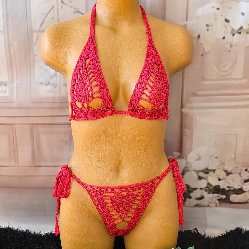 K&ouml;nnte beinhalten: Ein leuchtend pinkfarbenes H&auml;kel-Bikini-Set bestehend aus einem Neckholder-Triangel-Oberteil mit komplexen Lochmustern und einem passenden Bikini-Unterteil mit seitlichen Bindeb&auml;ndern, gefertigt aus Garn mit einem detaillierten geometrischen Spitzenmuster.