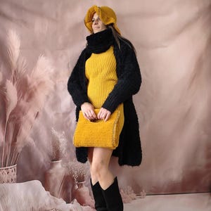 K&ouml;nnte beinhalten: Eine Frau tr&auml;gt ein senfgelbes Kleid, eine schwarze Strickjacke und schwarze Stiefel. Sie tr&auml;gt auch einen passenden senfgelben Hut und eine senfgelbe Handtasche. Das Outfit ist vor einem weichen rosa Hintergrund.