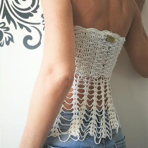 May include: A white strapless crochet top featuring an intricate openwork pattern with a scalloped design. The garment includes a small rectangular brown tag with the text Martima Hand Made attached to the upper right side, worn over blue denim jeans.