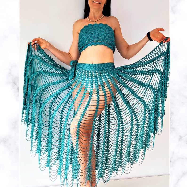May include: A teal crochet two-piece set consisting of a strapless tube top with a scalloped edge and a matching floor-length skirt. The skirt features an open, web-like mesh pattern with long, dangling vertical strands and a tie-side waist closure.
