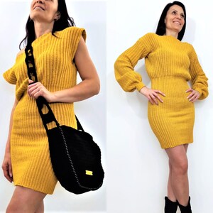 K&ouml;nnte beinhalten: Eine Frau tr&auml;gt ein senfgelbes Strickkleid und eine schwarze Umh&auml;ngetasche. Das Kleid hat eine gerippte Textur und eine figurbetonte Silhouette. Die Tasche ist gro&szlig; und rund mit einem dekorativen Riemen. Sie tr&auml;gt auch schwarze Stiefel.
