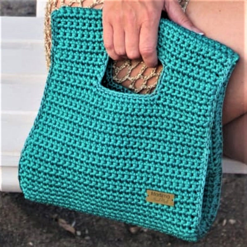 May include: A teal crochet handbag featuring a square cutout handle and a rectangular gold-toned metal tag with the text Martima Hand Made. The bag is constructed with a dense, textured stitch pattern and includes a gold chain strap detail.