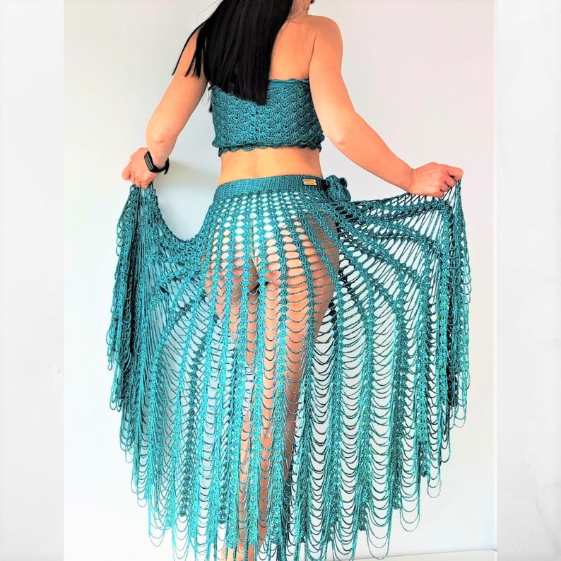 May include: A teal crochet two-piece set consisting of a strapless tube top and a long, sheer, open-weave skirt. The skirt features a wide waistband and a dramatic, flowing design with long, dangling vertical strands that create a fringe effect.