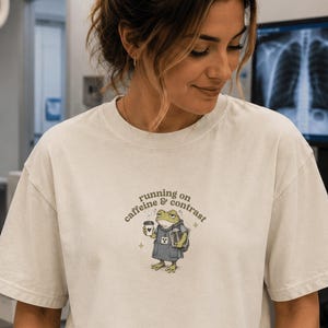 May include: A beige short-sleeved t-shirt featuring a graphic of a frog wearing a medical scrub top, holding a coffee cup and a medical folder. The text above the frog reads "running on caffeine & contrast" in a dark brown, curved font.