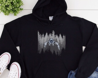 Peut inclure: Sweat-shirt à capuche noir avec un imprimé graphique de Bigfoot dans une forêt. Le graphique est en nuances de gris et de blanc.