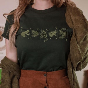 Pu&ograve; includere: Una maglietta verde scuro con un design circolare di cinque rane. La maglietta &egrave; indossata da una persona con capelli lunghi castani e un cardigan marrone. La persona indossa una gonna marrone con bottoni sul davanti.