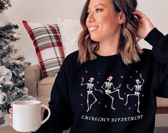 Op de afbeelding: Zwarte sweatshirt met een ontwerp van drie dansende skeletten met kerstmutsen en de tekst "EMERGENCY DEPARTMENT". De achtergrond toont een versierde kerstboom en een cadeauverpakking.