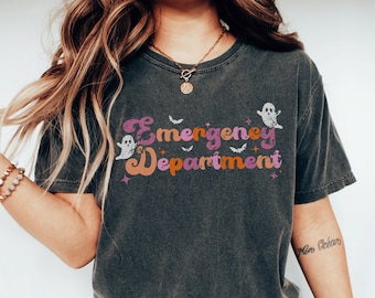 Op de afbeelding: Donkergrijs t-shirt met de tekst "Emergency Department" in een retro lettertype, met geesten en vleermuizen. De tekst is in tinten paars, oranje en roze. Het shirt heeft een casual stijl.