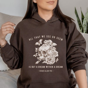 Può includere: Una felpa con cappuccio marrone con una grafica di teschio e fiori e il testo "ALL THAT WE SEE OR SEEM IS BUT A DREAM WITHIN A DREAM - EDGAR ALLAN POE -"