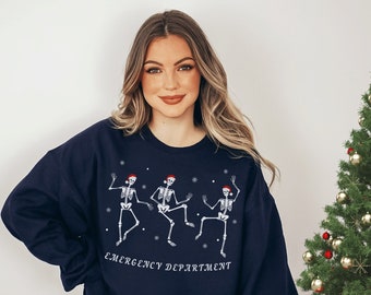 Op de afbeelding: Marineblauw sweatshirt met drie dansende skeletfiguren met kerstmutsen en sneeuwvlokken. De tekst "Emergency Department" staat eronder. Een feestelijk kledingstuk met een vakantiethema.