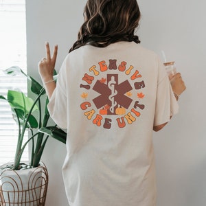 Peut inclure: T-shirt cr&egrave;me avec un motif circulaire au dos. Le motif indique "INTENSIVE CARE UNIT" en lettres orange et marron, avec un symbole m&eacute;dical marron, des citrouilles et des feuilles d'automne.