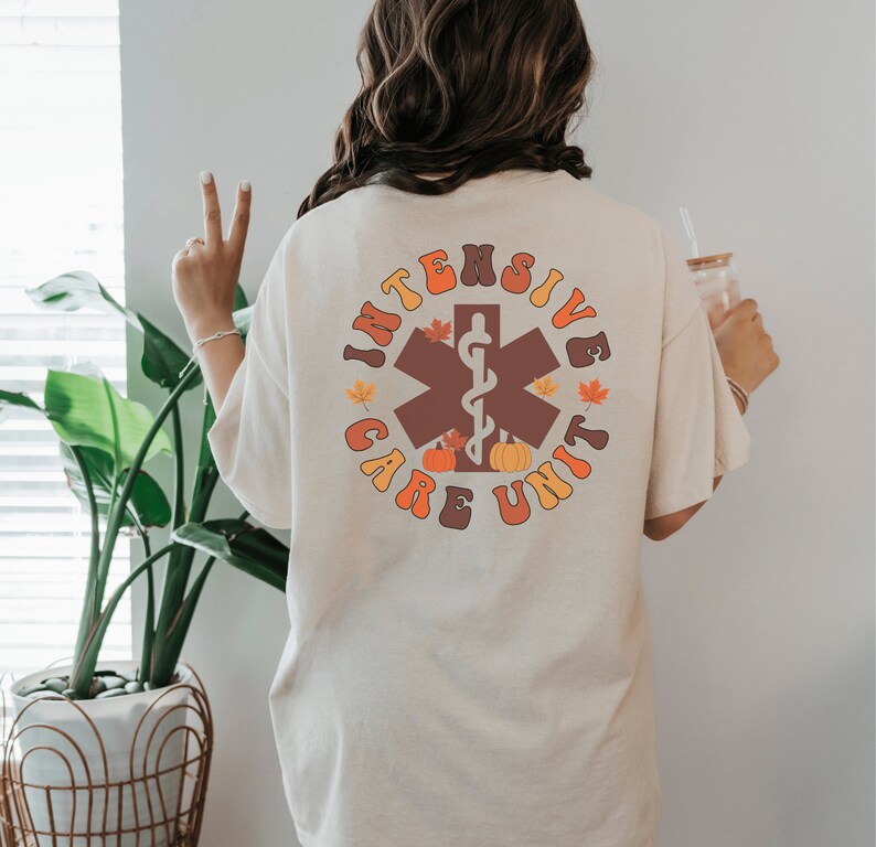Peut inclure: T-shirt cr&egrave;me avec un motif circulaire au dos. Le motif indique "INTENSIVE CARE UNIT" en lettres orange et marron, avec un symbole m&eacute;dical marron, des citrouilles et des feuilles d'automne.