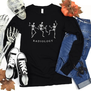 Op de afbeelding: Zwart shirt met lange mouwen met een afbeelding van drie dansende skeletten en de tekst "RADIOLOGY".