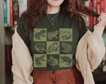 May include: Dark green t-shirt featuring a grid of nine frog illustrations in a vintage style. The frogs are in various poses and are in a lighter green color. The shirt is paired with a brown skirt and a cream-colored cardigan.
