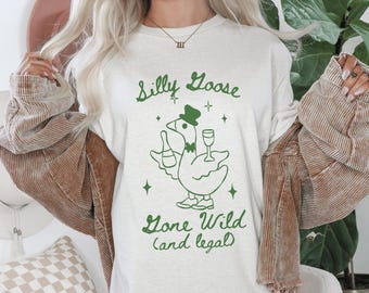 Puede incluir: Camiseta color crema con un diseño verde de un ganso con sombrero, botella y copa de vino. El texto dice "Silly Goose Gone Wild (and legal)".