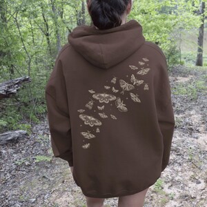 May include: A brown hooded sweatshirt featuring a cream-colored moth and butterfly design on the back. The design includes various sizes and shapes of moths and butterflies, creating a whimsical and nature-inspired aesthetic. The sweatshirt is a solid brown color.