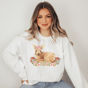 May include: White sweatshirt featuring a fawn with a floral crown, lying amidst flowers and mushrooms. The fawn is light brown with white spots. The flowers are pink and the mushrooms are red and white. The sweatshirt is a casual, comfortable style.