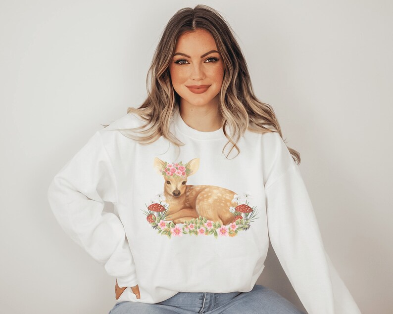 May include: White sweatshirt featuring a fawn with a floral crown, lying amidst flowers and mushrooms. The fawn is light brown with white spots. The flowers are pink and the mushrooms are red and white. The sweatshirt is a casual, comfortable style.