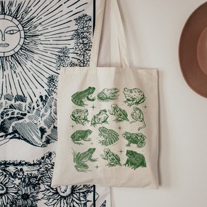 May include: A cream tote bag with a green print of different frog illustrations. The frogs are drawn in a vintage style, with detailed outlines and shading. The bag is suspended against a white wall, with a decorative tapestry and a hat in the background.