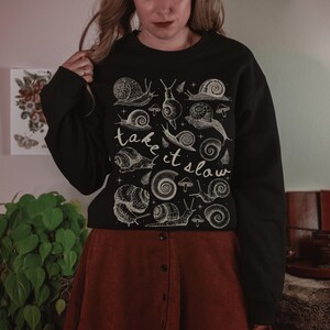 Puede incluir: Sudadera negra con una ilustraci&oacute;n blanca de caracoles y setas. El texto "take it slow" est&aacute; escrito en cursiva. La sudadera se combina con una falda color &oacute;xido.