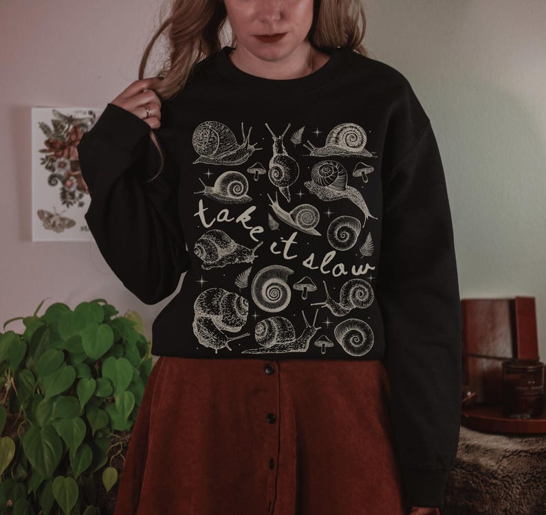 Puede incluir: Sudadera negra con una ilustraci&oacute;n blanca de caracoles y setas. El texto "take it slow" est&aacute; escrito en cursiva. La sudadera se combina con una falda color &oacute;xido.