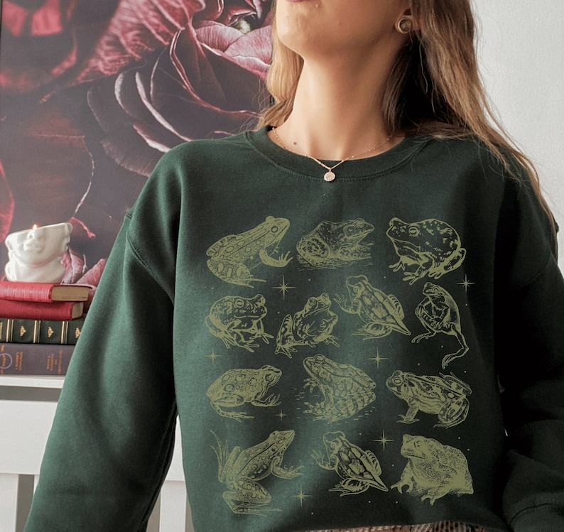 May include: A dark green sweatshirt with a graphic print of frogs in various poses. The frogs are outlined in gold and are set against a dark green background.