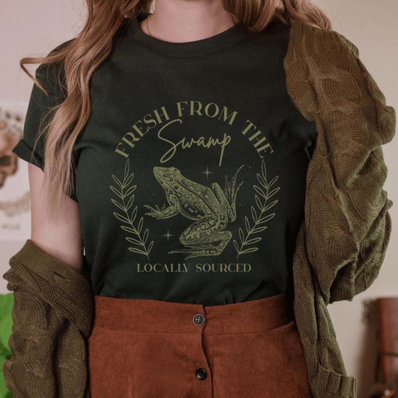 May include: Dark green t-shirt with a frog graphic and the text "FRESH FROM THE SWAMP LOCALLY SOURCED". The frog is green and detailed, surrounded by a laurel wreath design. The shirt is a short-sleeved crew neck.