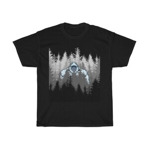 Pu&ograve; includere: Maglietta nera con una grafica di un Bigfoot bianco in piedi in una foresta di alberi scuri.