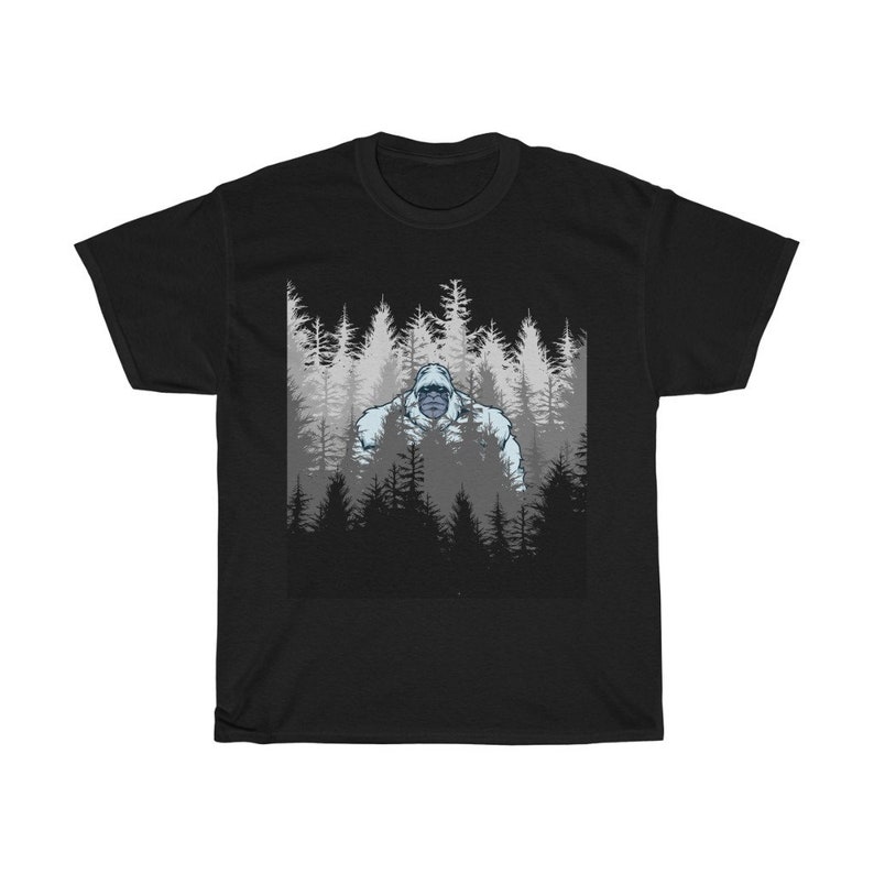 Pu&ograve; includere: Maglietta nera con una grafica di un Bigfoot bianco in piedi in una foresta di alberi scuri.