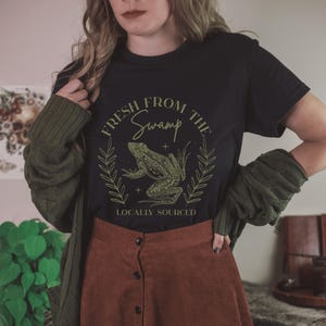 May include: Black t-shirt with a green frog graphic inside a laurel wreath. The text reads "FRESH FROM THE SWAMP" above the frog and "LOCALLY SOURCED" below. The shirt is paired with a brown skirt and a green cardigan.
