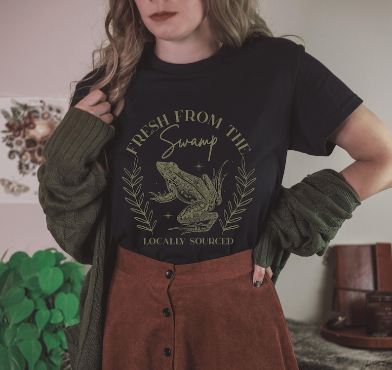 May include: Black t-shirt with a green frog graphic inside a laurel wreath. The text reads "FRESH FROM THE SWAMP" above the frog and "LOCALLY SOURCED" below. The shirt is paired with a brown skirt and a green cardigan.
