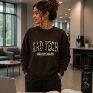 May include: A dark brown crewneck sweatshirt featuring the text RAD TECH in a white leopard print pattern, with the words IN PROGRESS printed in a white rectangular outline below. The sweatshirt has long sleeves and a ribbed collar and cuffs.