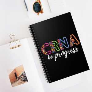 Puede incluir: Una libreta espiral negra con el texto "CRNA in progress" en letras de colores con diseños florales.