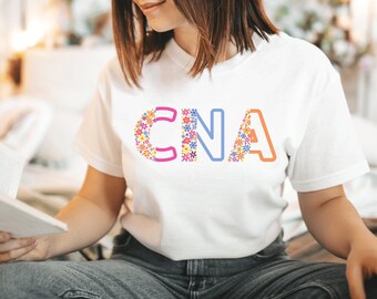 May include: A white short-sleeved t-shirt featuring the letters CNA printed on the chest. The letters are stylized with a colorful floral pattern consisting of small red, yellow, blue, and purple flowers, combined with solid pink, blue, and orange outlines.