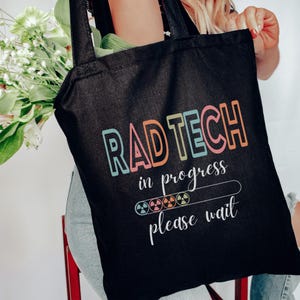 May include: Black tote bag with the words "RAD TECH" in colorful letters. Below it, the text "in progress" and "please wait" are displayed. The bag features a graphic of a loading bar with radiation symbols.