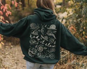 May include: Dark green hoodie with a white snail and mushroom graphic on the back. The text "take it slow" is also printed on the back. The hoodie has a hood and is made of a soft material.