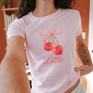 May include: Pink t-shirt with a graphic of two red cherries tied together with a pink ribbon. The text "Little" is printed below the cherries.