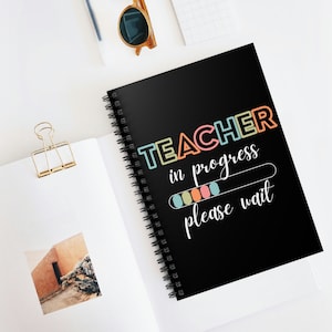 Puede incluir: Un cuaderno negro con espiral con el texto "TEACHER in progress please wait" en letras coloridas. El cuaderno está abierto sobre una superficie blanca, con un clip dorado y gafas de sol visibles.