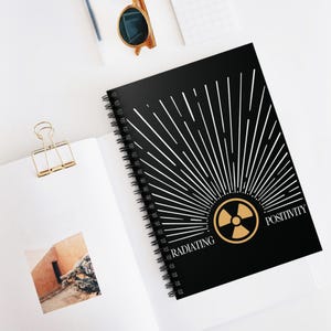 Op de afbeelding: Zwart spiraalgebonden notitieboek met een goud en wit ontwerp. De omslag heeft een sunburst-ontwerp met de woorden "Radiating Positivity" en een gouden stralingssymbool. Het notitieboek is open op een wit oppervlak.