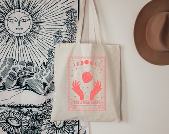 Puede incluir: Una bolsa de lona blanca con un diseño rosa que presenta una fresa, manos y una luna creciente. El texto "THE STRAWBERRY" está impreso en la bolsa.