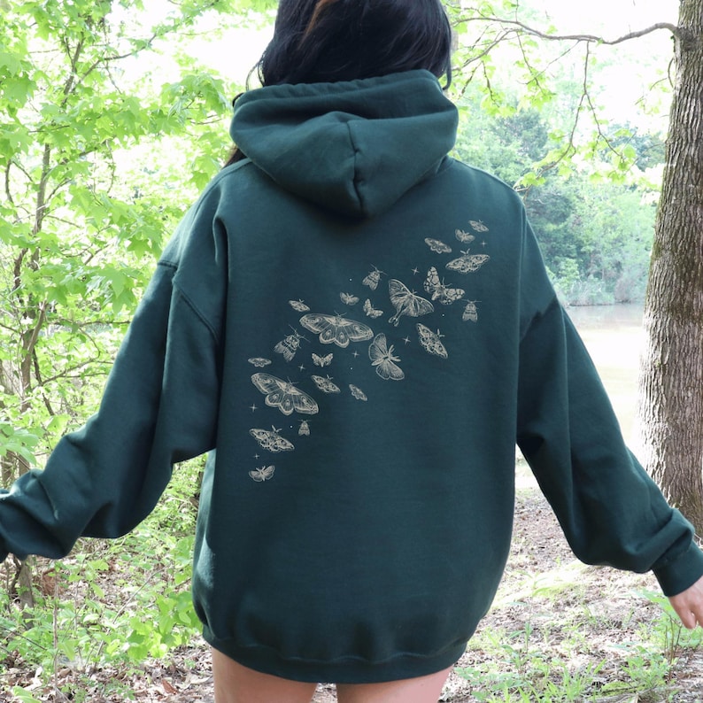 May include: A dark green hoodie with a hood and long sleeves. The back of the hoodie features a design of several moths in various sizes and orientations, in a light tan color. The hoodie is made of a soft material.